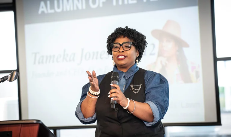 Tameka Jones at Saint Paul College alumni awards ceremony, presenting with a microphone in front of “Alumni of the Year” slide.