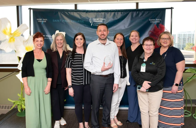 Group photo of Saint Paul College staff standing in front of branded Saint Paul College backdrop at an indoor event, featuring nine employees posing together with a glass award in a bright office setting with large windows and decorative flowers.