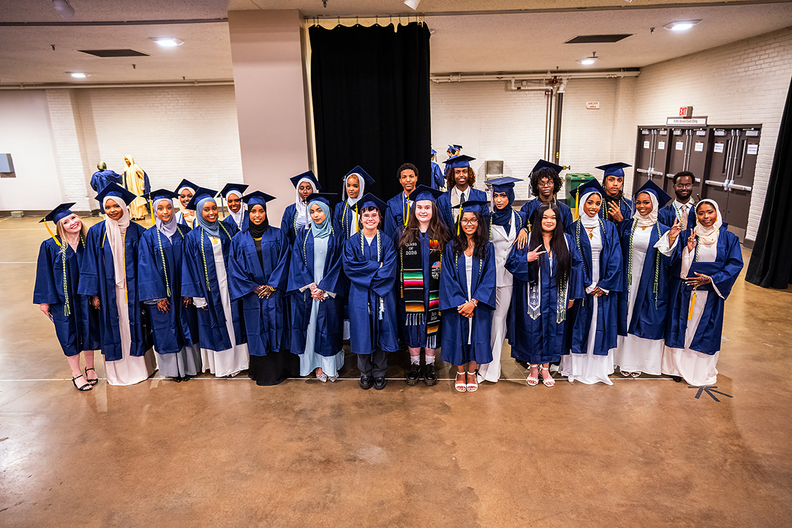 Group of Saint Paul College graduates in blue caps and gowns pose together in a large indoor hallway before the ceremony.