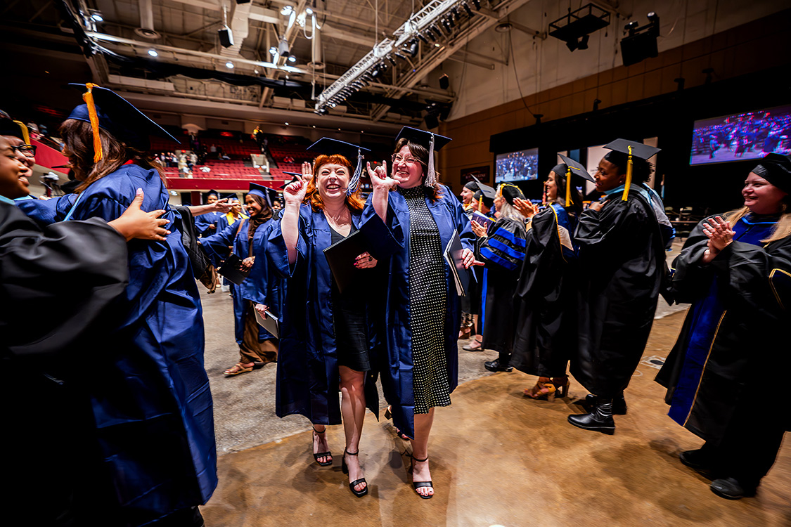 Graduates in navy caps and gowns walk through an auditorium aisle while others on both sides clap and reach out, with stage screens and seating visible in the background. (Spring 2026 graduation ceremony)