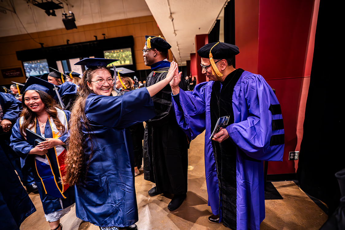 Saint Paul College graduate in a blue cap and gown high-fives a faculty member in academic regalia backstage at graduation, with other graduates waiting nearby.