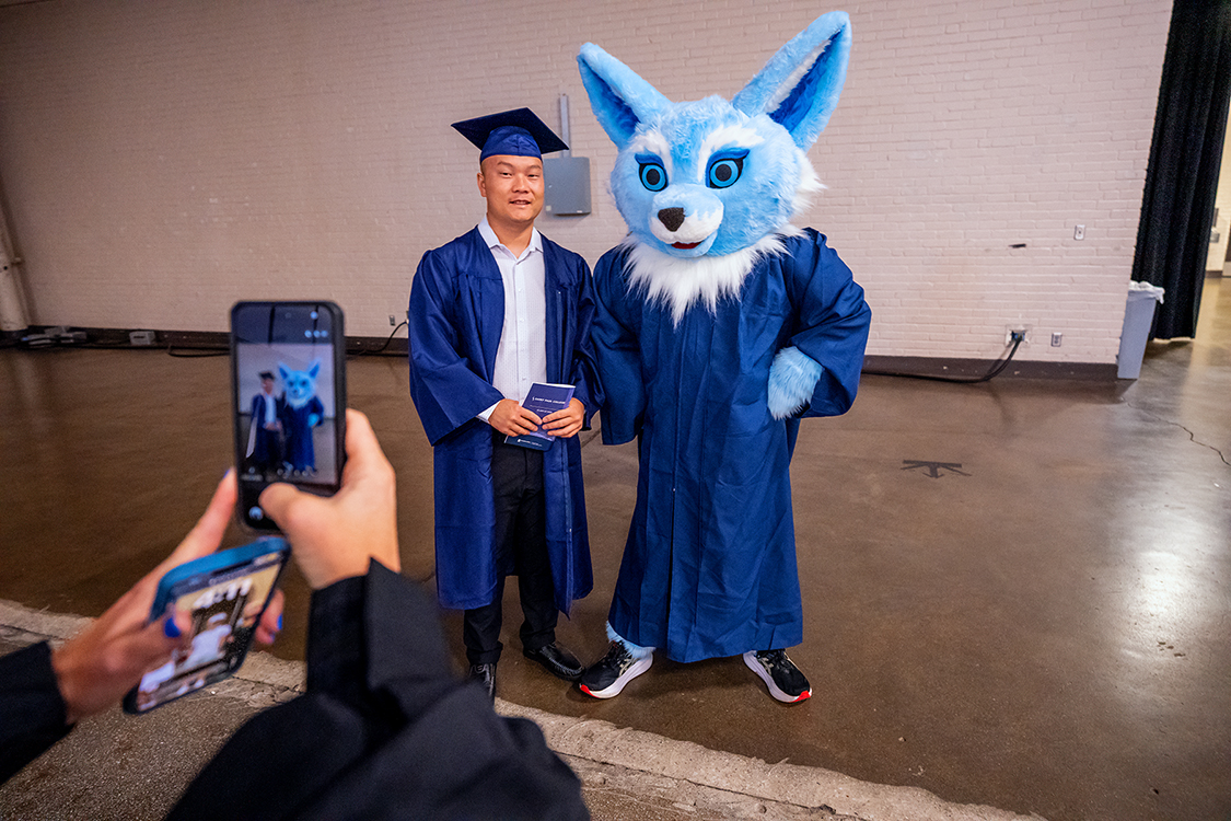 Saint Paul College graduate in a blue cap and gown poses for a photo with Sky the SPC mascot, a blue fox in a graduation gown, while someone takes the picture on a phone.