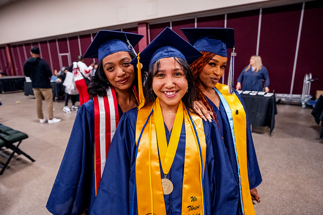 Three Saint Paul College graduates in blue caps and gowns pose together indoors, wearing honor cords, stoles, and a medal before the graduation ceremony.