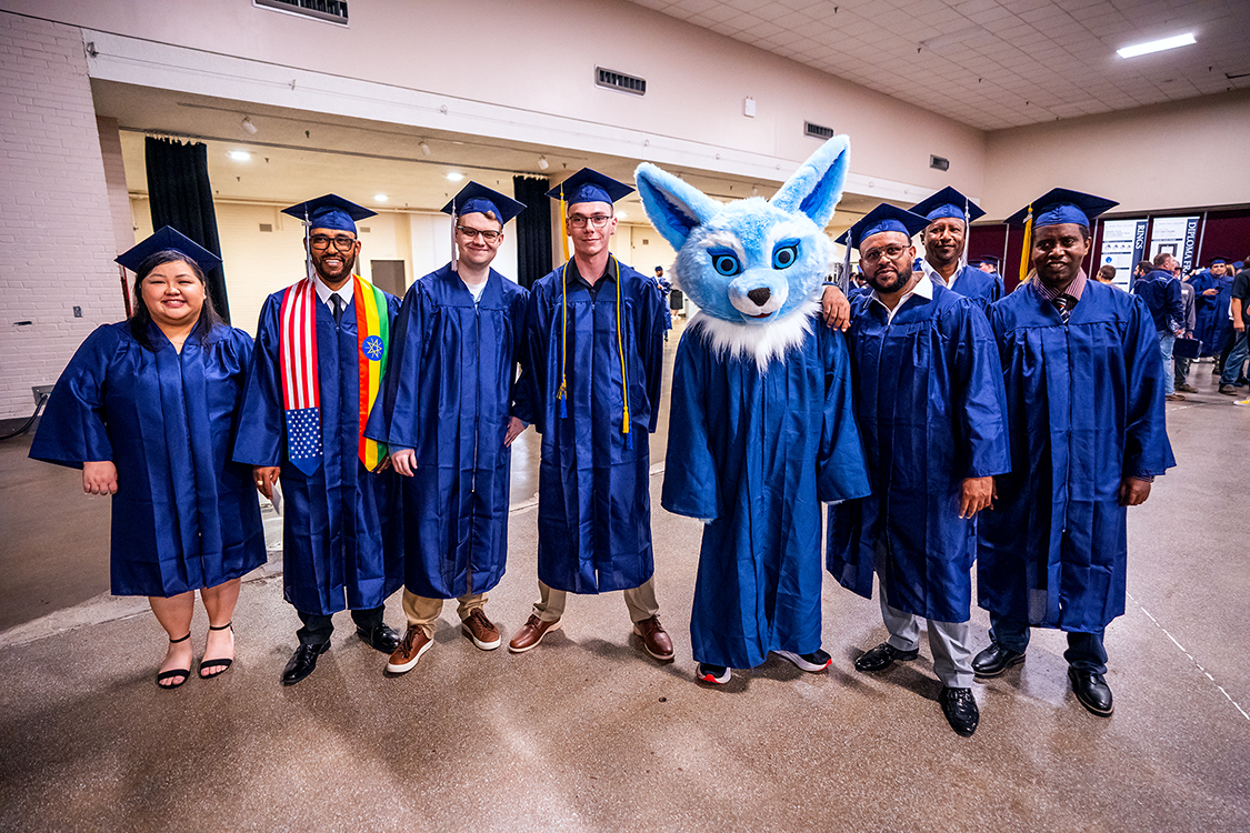 A group of Saint Paul College graduates in blue caps and gowns pose for a photo with Sky the SPC mascot in a blue graduation gown.