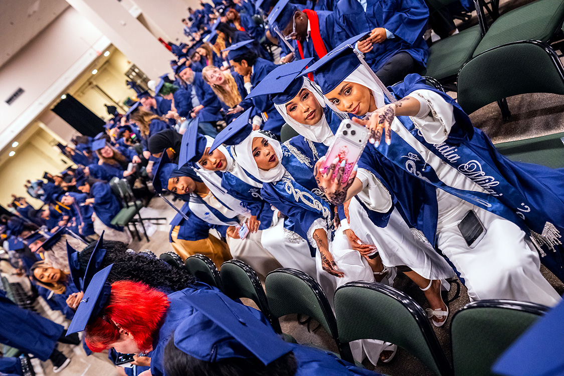 A group of seated graduates in caps and gowns take a selfie on a smartphone while surrounded by rows of other graduates in an auditorium. (Spring 2026 graduation ceremony)