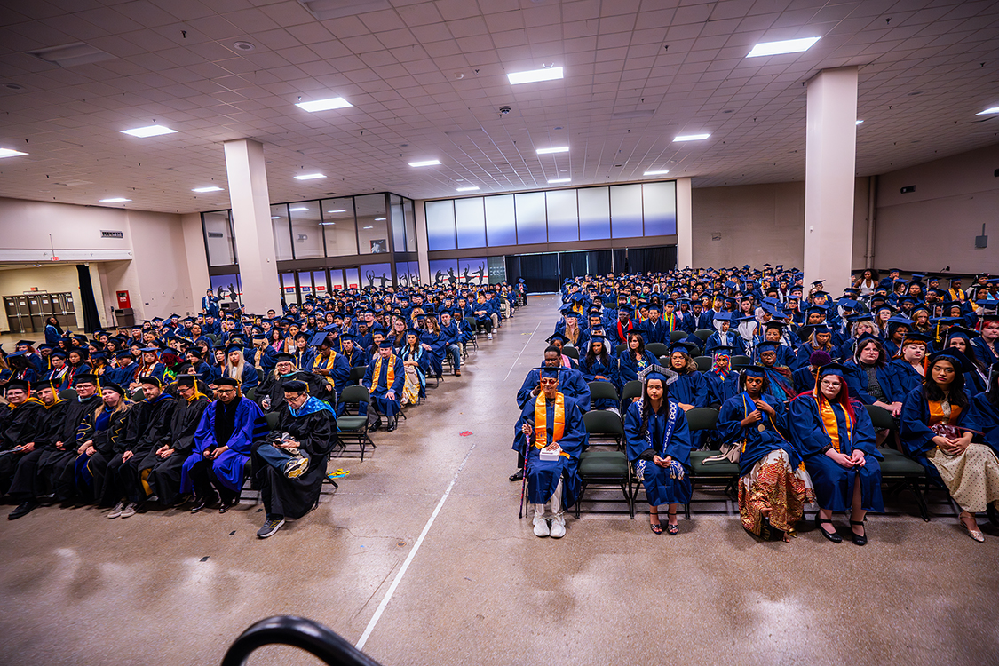 Hundreds of Saint Paul College graduates in blue caps and gowns sit in rows of chairs inside a large indoor venue before the graduation ceremony begins.