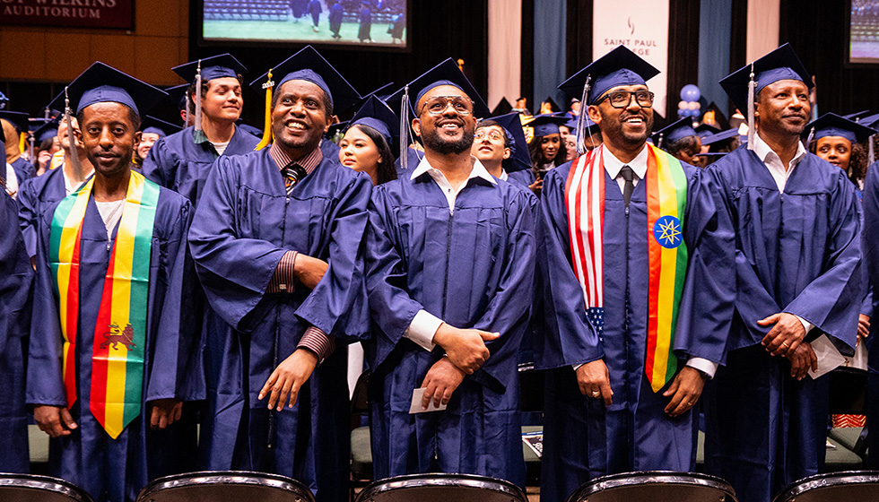 A group of graduates in navy-blue caps and gowns stand together in an auditorium, some wearing colorful striped stoles, with a stage and “Saint Paul” signage visible in the background. (Spring 2026 graduation ceremony)