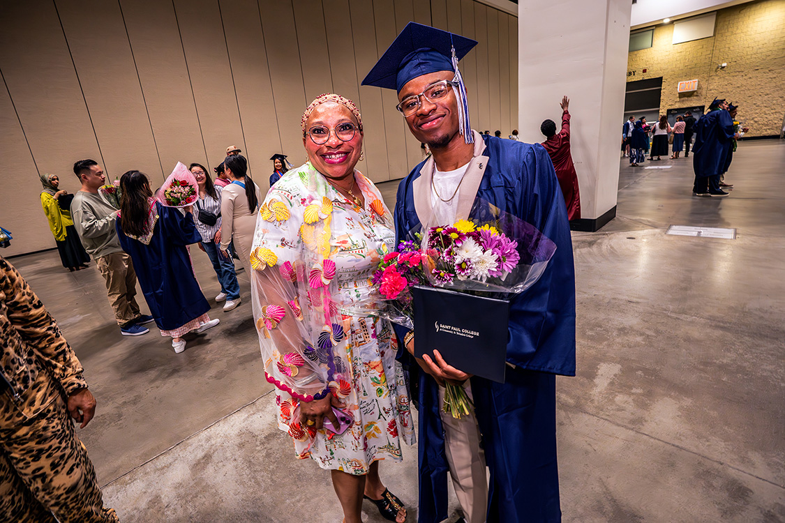 Graduate in a navy cap and gown holds a diploma folder and bouquet while posing with a person holding flowers in a large indoor venue. (Spring 2026 graduation ceremony)