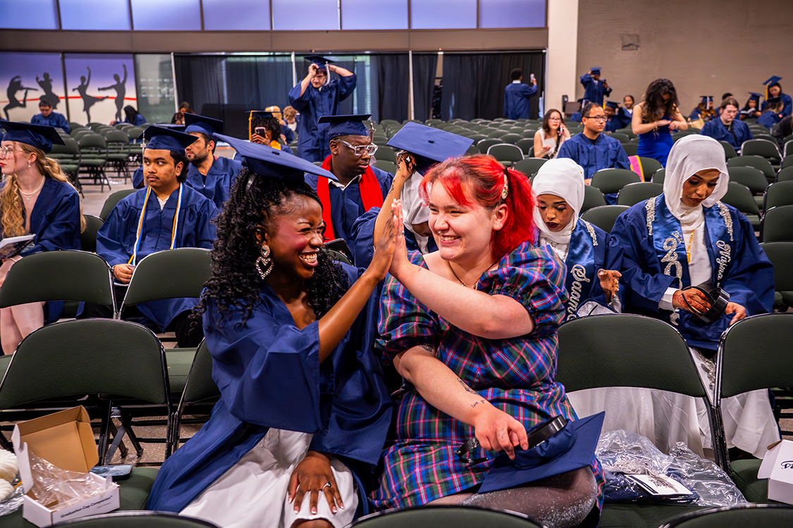 A Saint Paul College graduate in a blue gown sits among rows of chairs and high-fives a person seated beside them before the graduation ceremony, with other graduates in blue caps and gowns in the background.