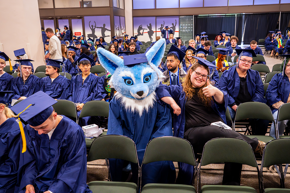 Sky the SPC mascot, wearing a blue cap and gown, sits among Saint Paul College graduates in rows of chairs before the graduation ceremony while a person beside the mascot gives a thumbs-up.