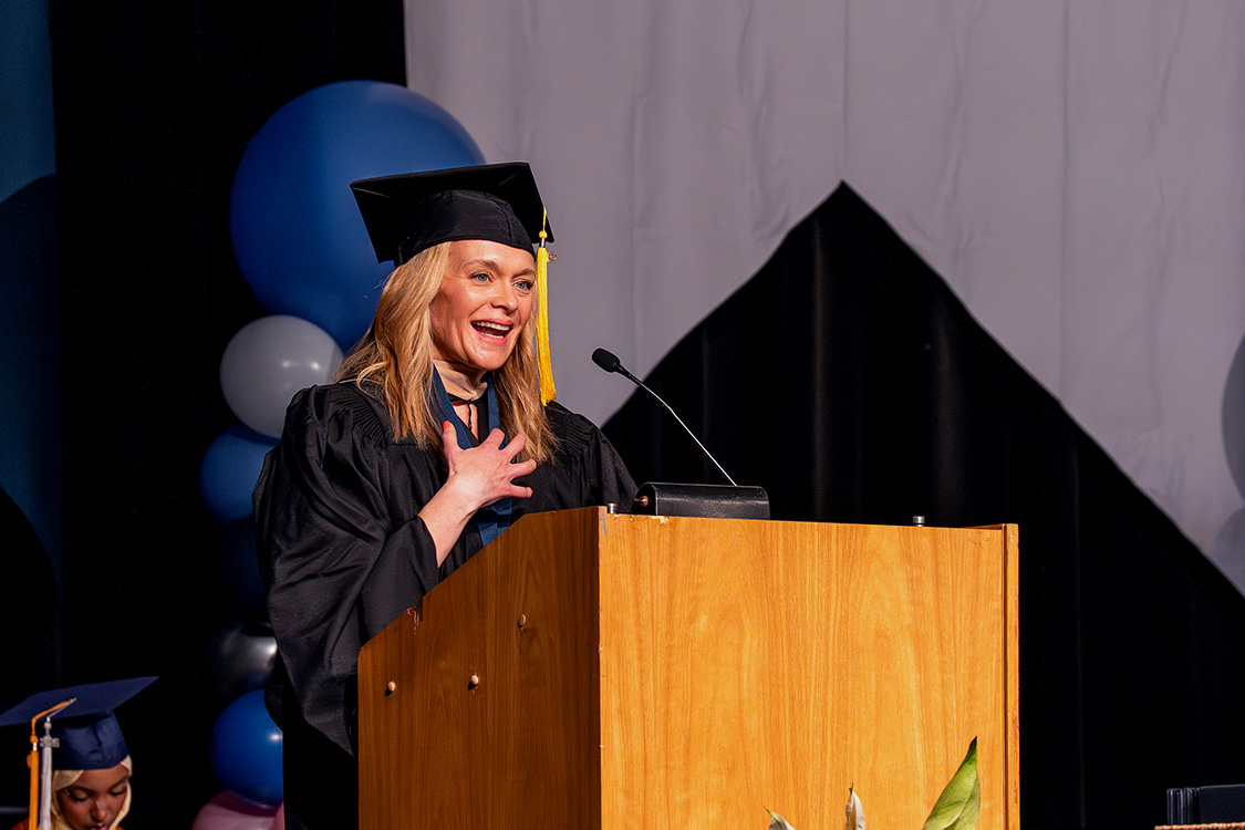 Mindy Travers in a graduation cap and gown stands at a podium on stage with balloons and curtains in the background for the Class of 2026 graduation. (Spring 2026 graduation ceremony)