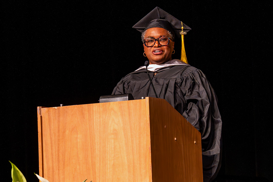 Wendy Roberson in academic regalia and cap delivers remarks at a podium against a dark stage backdrop for the Class of 2026 graduation. (Spring 2026 graduation ceremony)