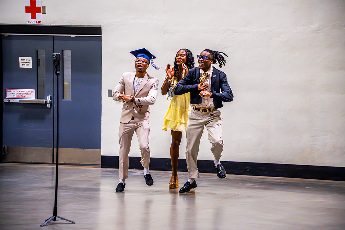 A Saint Paul College graduate wearing a blue cap dance with two formally dressed people in an open indoor hallway during graduation.