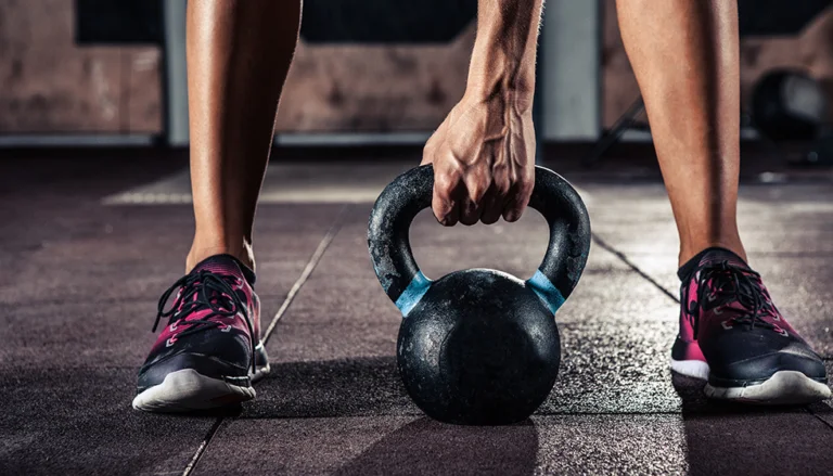 Close-up of a person lifting a black kettlebell from a gym floor, wearing athletic shoes, highlighting strength training and functional fitness.