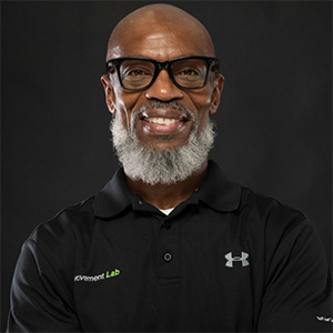 Portrait of Charles “Coach Chuck” Turner wearing glasses and a black polo shirt with a “Movement Lab” logo, standing against a dark background with arms crossed.