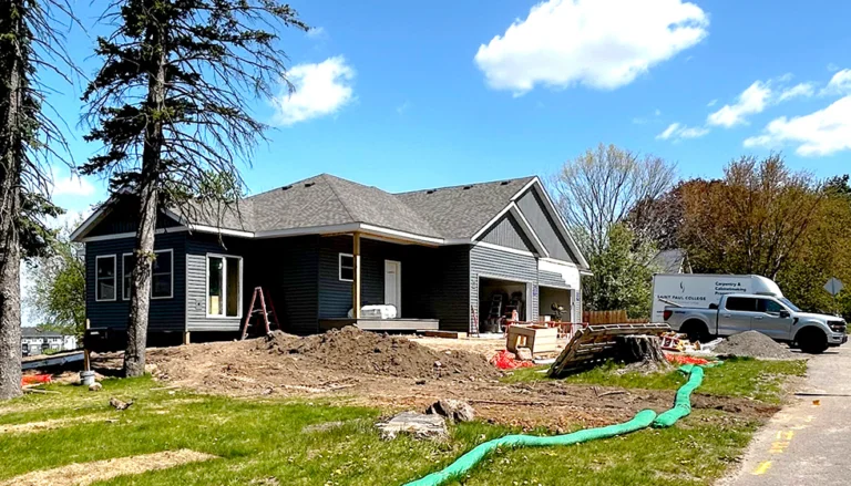 Newly constructed duplex on a residential lot with gray siding, attached garage, and landscaping in progress, with construction materials and a Saint Paul College work truck visible on site.