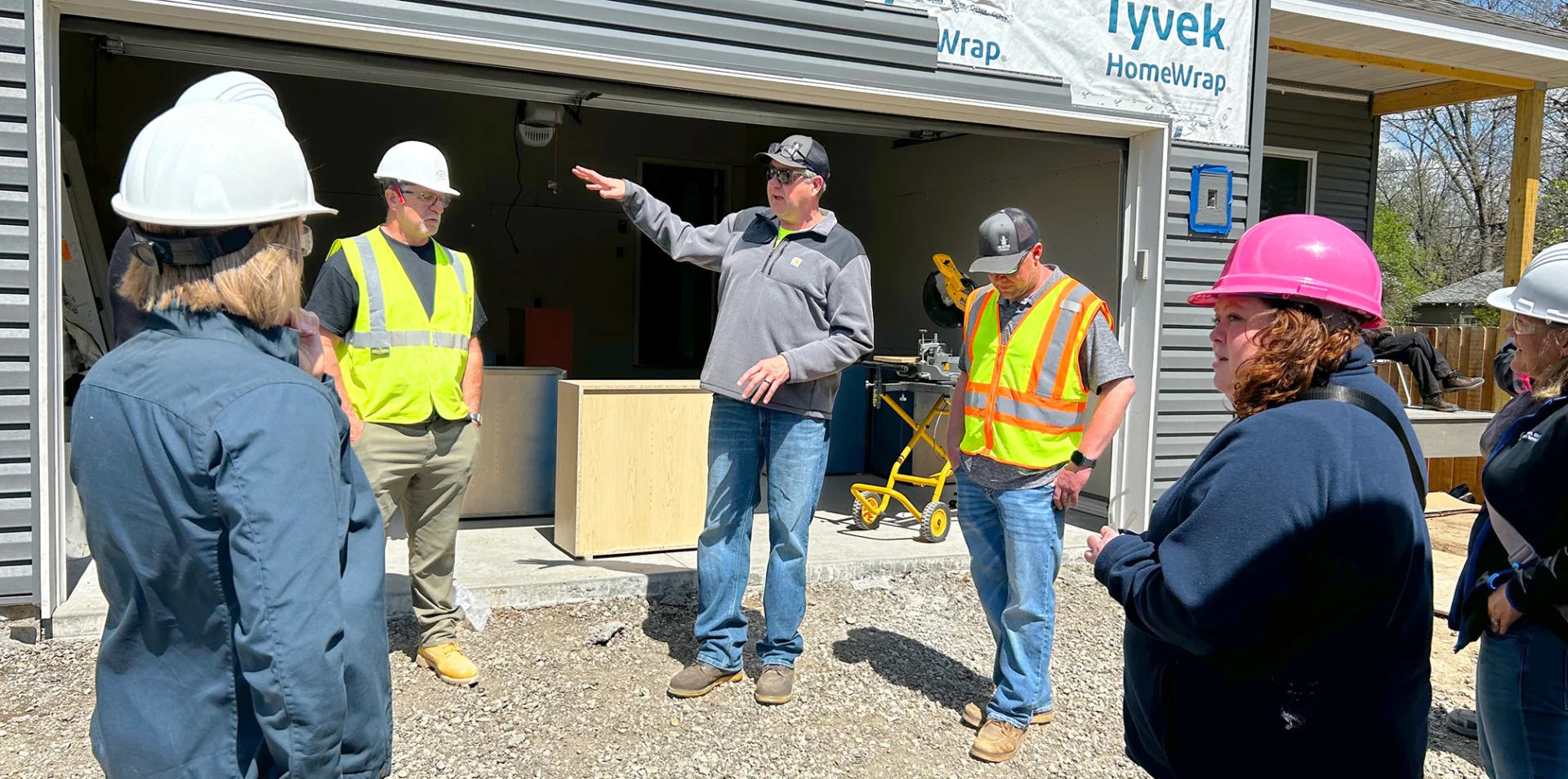 Group of construction students and instructors wearing hard hats and safety vests gathered outside a garage, listening to instructions during a hands-on building project at the duplex site.