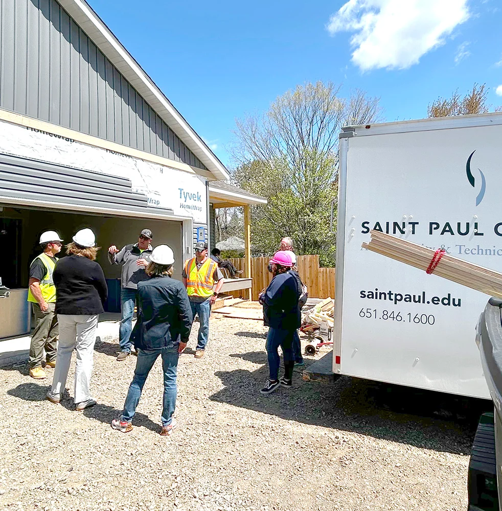 Students and instructors stand near the duplex under construction alongside a Saint Paul College trailer, discussing progress on the residential build project in North Saint Paul.