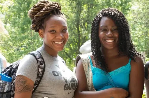 Two young women smiling outdoors on a sunny day, standing side by side with backpacks and tote bags.