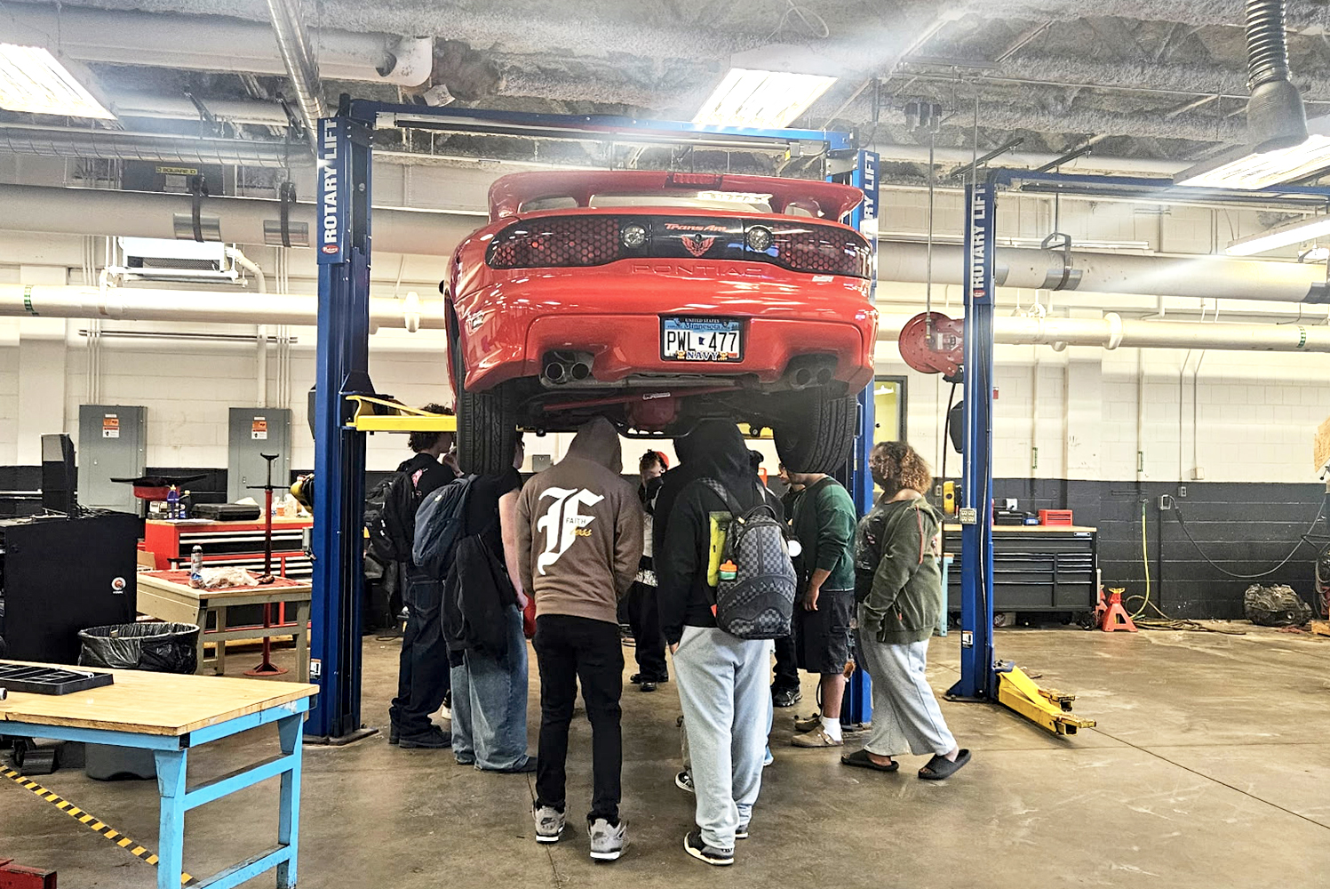 Group of high school students under a red car on a lift being shown what an automotive technician will work on.