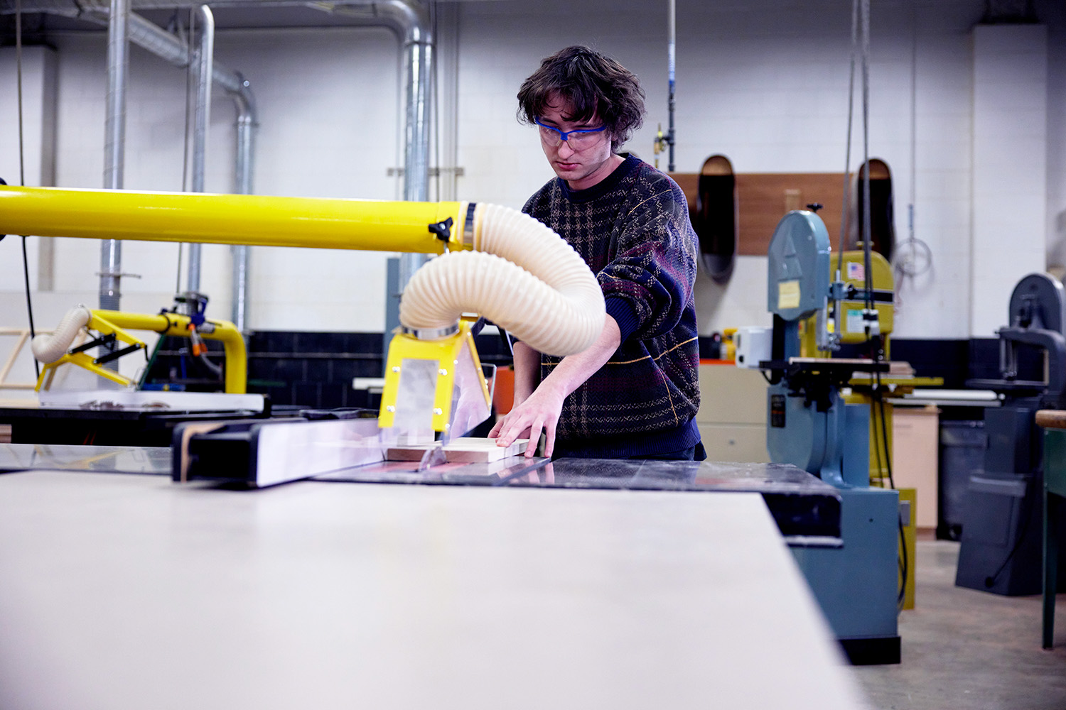 A student wearing safety glasses operates a woodworking machine in a shop classroom. The student guides a piece of wood across the machine’s surface while a large yellow dust-collection hose arches overhead. Additional woodworking equipment is visible throughout the workshop.