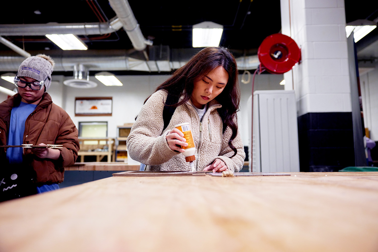 Two students work on a woodworking project at a large workbench. In the foreground, one student applies glue to a wood piece, while another student stands nearby holding project materials. The workshop contains tools, workstations, and safety equipment in the background.