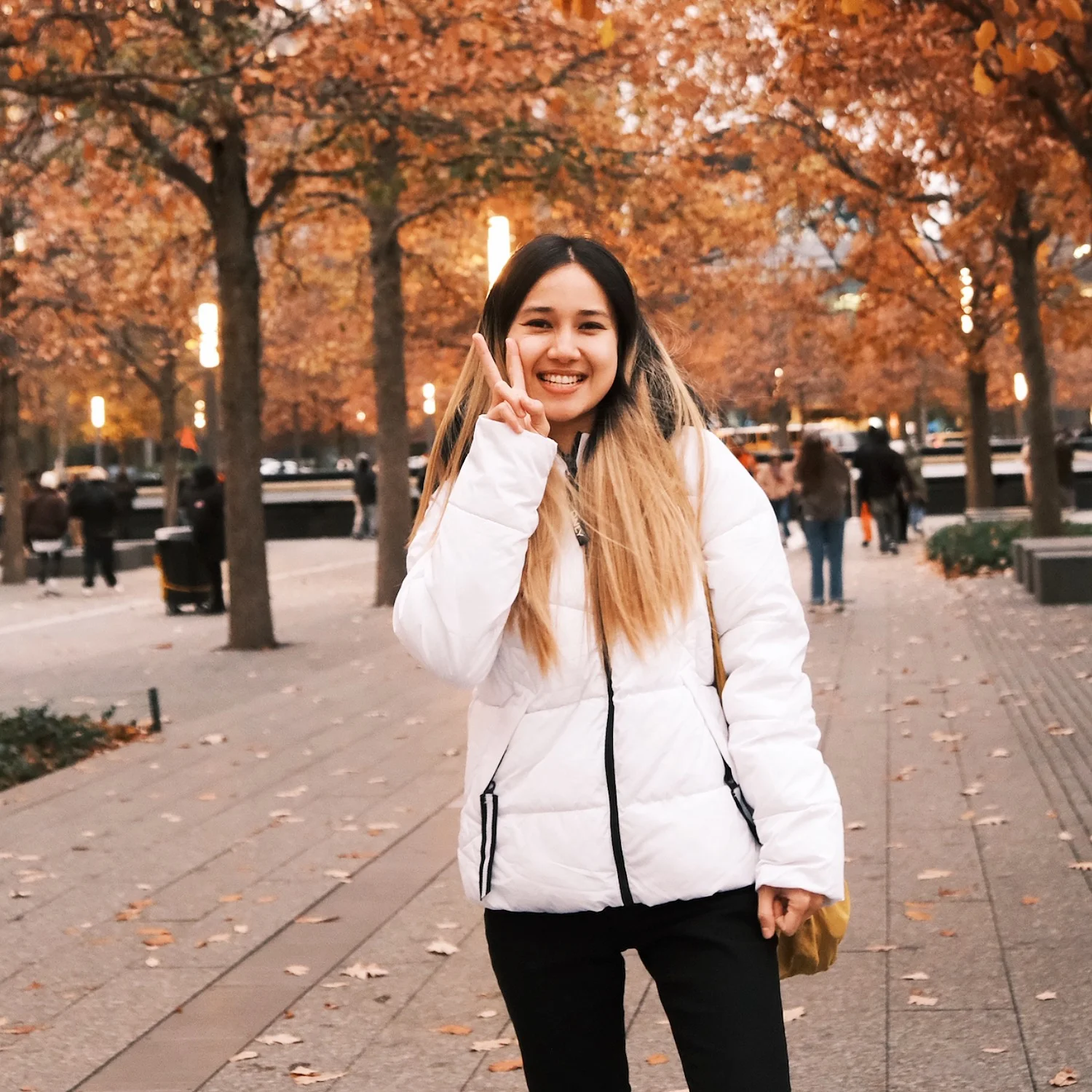 Eaint Phu Myint stands on a tree-lined walkway during autumn, wearing a white puffer jacket and carrying a shoulder bag. She raises a peace sign toward the camera, with orange fall foliage, pathway lights, and pedestrians visible in the background.