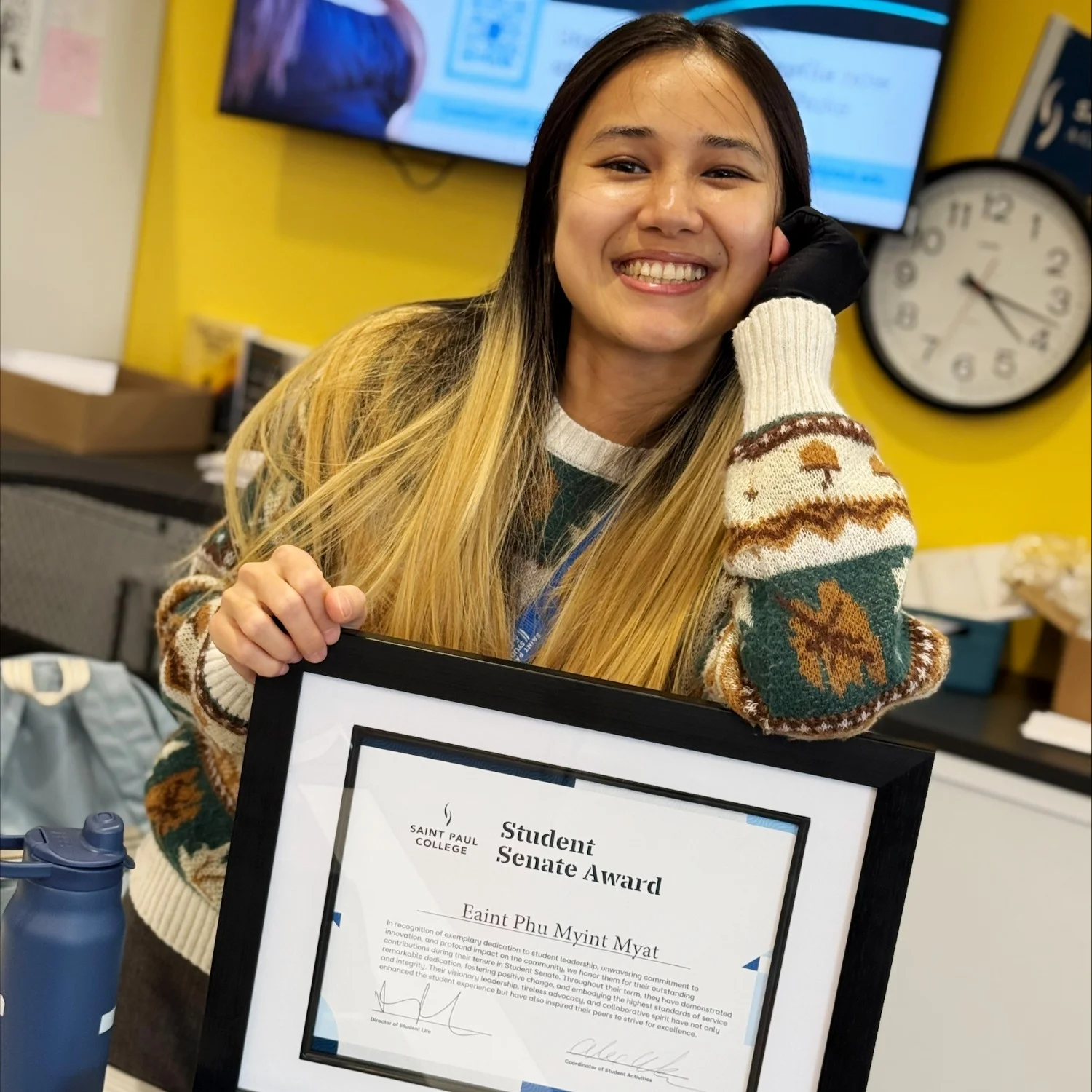 Eaint Phu Myint holds a framed Saint Paul College Student Senate Award certificate while seated indoors. The photo shows Eaint Phu Myint wearing a patterned sweater and a campus lanyard, with the framed award prominently displayed in the foreground. A yellow wall, television screen, and wall clock are visible in the background.