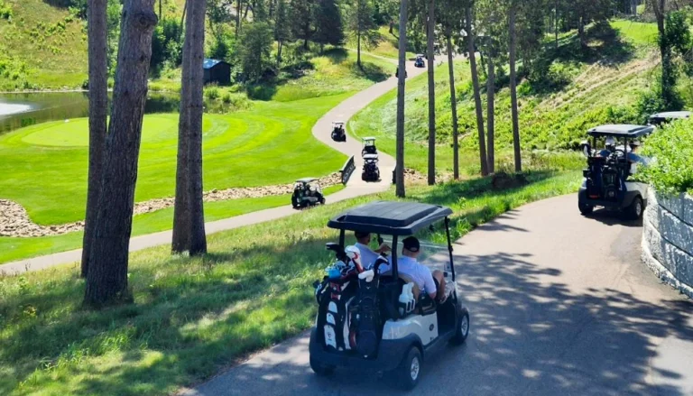 Golf carts travel along a winding paved path through a lush, tree-lined golf course on a sunny day. Bright green fairways and a small pond are visible to the left, while tall pine trees and rolling hills surround the course. Several golfers ride in carts along the path, with one cart in the foreground carrying golf bags.
