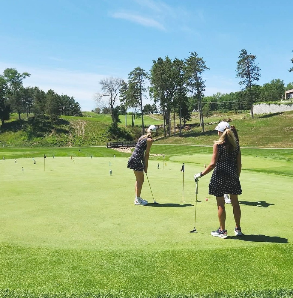 Two golfers practice putting on a large, manicured putting green at a scenic golf course. Multiple practice holes and markers are spread across the green, with rolling hills, trees, and a small bridge visible in the background under a clear blue sky.