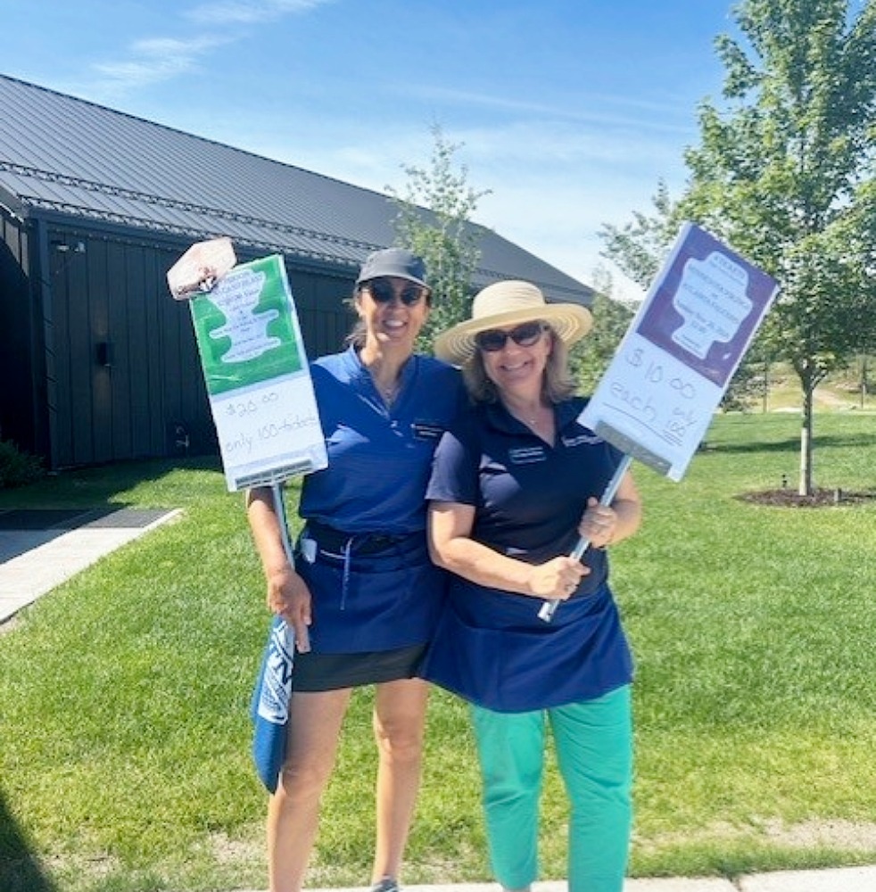 Two event volunteers stand on a grassy area outside a golf facility, holding promotional signs. Trees, a dark-colored building, and a sunny blue sky provide the backdrop.