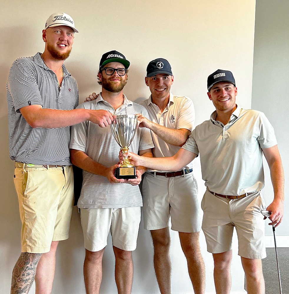 Four golfers pose indoors with a large trophy, standing side by side in golf attire. One golfer holds a golf club while the group displays the trophy against a plain wall background.