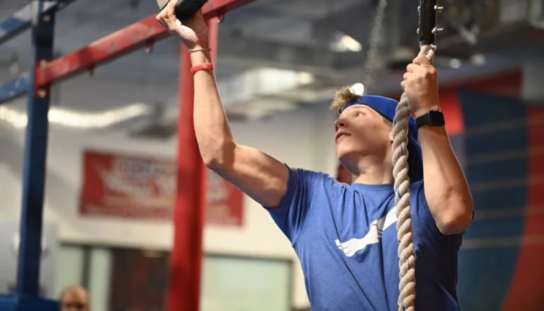 Person in a blue T-shirt and backward blue cap navigating an indoor ninja obstacle course. The person grips a hanging rope with one hand and reaches upward toward a suspended obstacle with the other. Red and blue metal obstacle structures frame the scene, and a smartwatch is visible on one wrist. The background is softly blurred, showing additional gym equipment and signage.