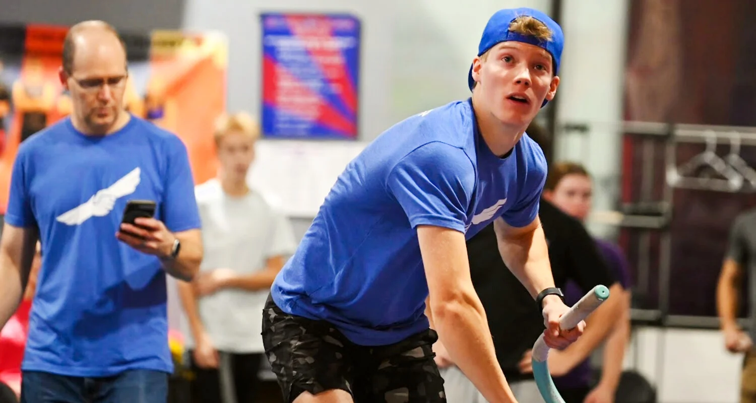 Person in a blue T-shirt and backward blue cap navigating an indoor ninja obstacle course. The person grips a hanging rope with one hand and reaches upward toward a suspended obstacle with the other. Red and blue metal obstacle structures frame the scene, and a smartwatch is visible on one wrist. The background is softly blurred, showing additional gym equipment and signage.