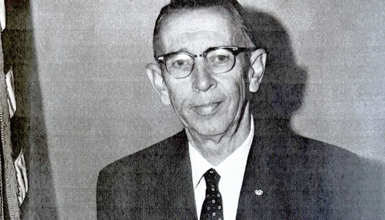 Black-and-white portrait photograph of a Harold Ostrem wearing glasses, a dark suit jacket, white dress shirt, and patterned tie, standing indoors against a plain wall. Part of a flag is visible along the left edge of the image. The photograph has a grainy, archival appearance.