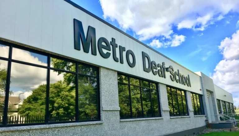 Exterior view of a modern commercial building with large windows and a white facade displaying the sign “Metro Deaf School.” Reflections of trees appear in the windows beneath a bright blue sky with scattered clouds.