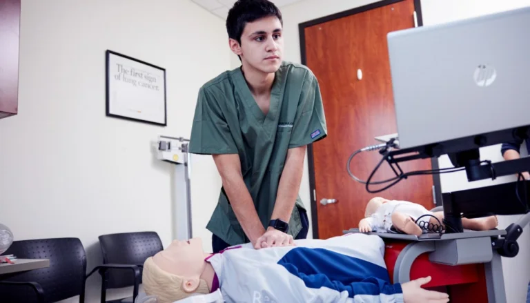 Person in green medical scrubs practices CPR on a medical training mannequin in a clinical simulation room. A computer workstation and infant mannequin are positioned nearby, with chairs, medical equipment, and a door visible in the background.