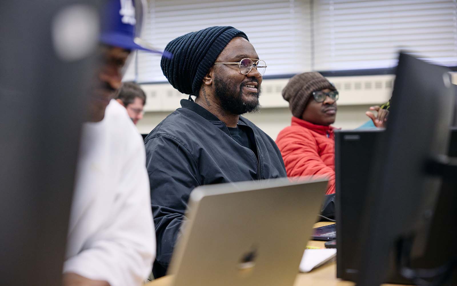 Students work at computers in a classroom computer lab, with one student in focus seated behind a laptop.