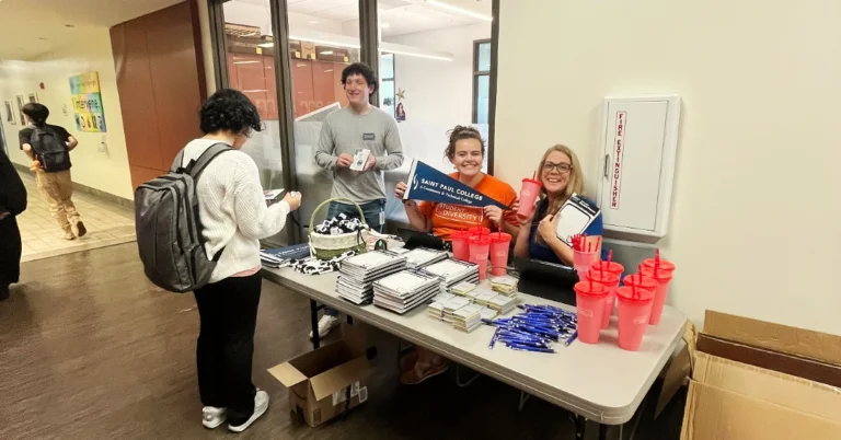 Saint Paul staff tabling with college swag with one student grabbing swag and one helping table.