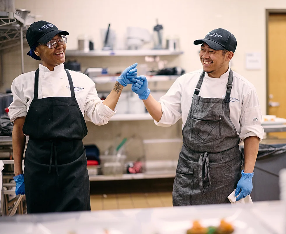 Two Saint Paul College culinary students in chef uniforms and gloves greet each other while working in a commercial teaching kitchen.