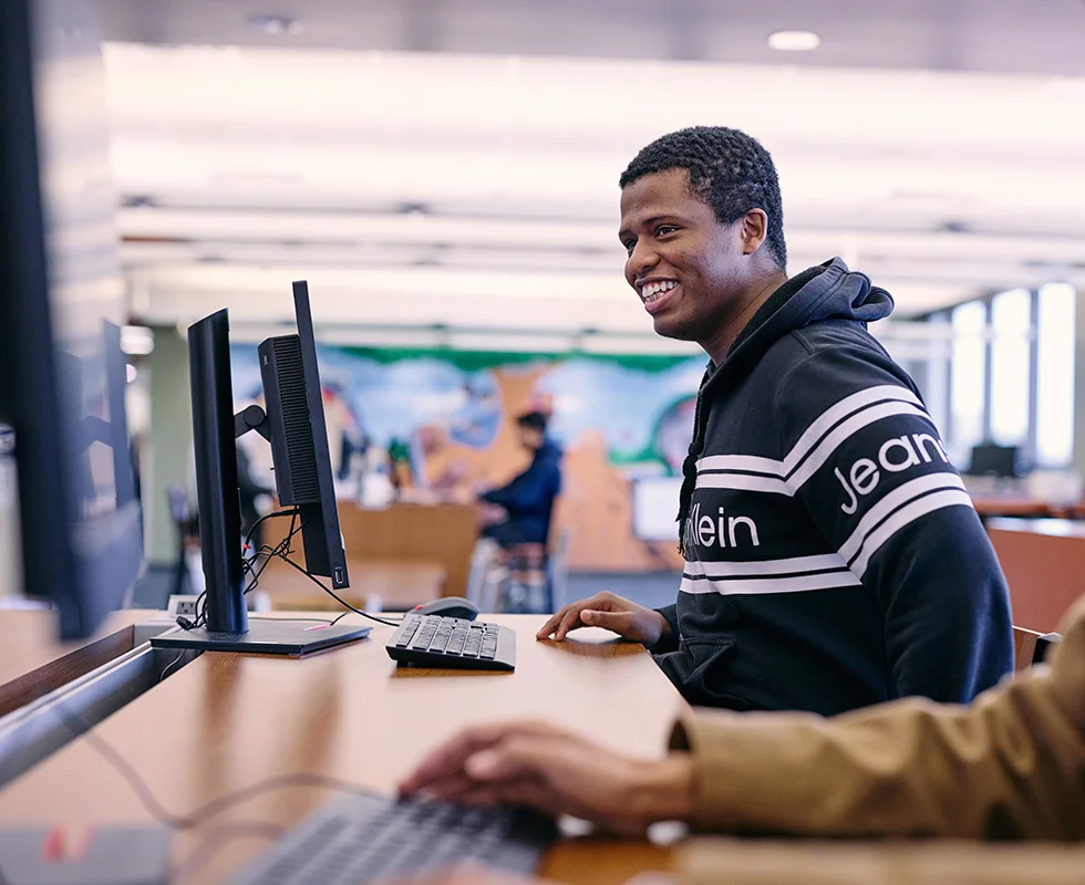Saint Paul College student uses a computer workstation while receiving assistance at a campus service desk.