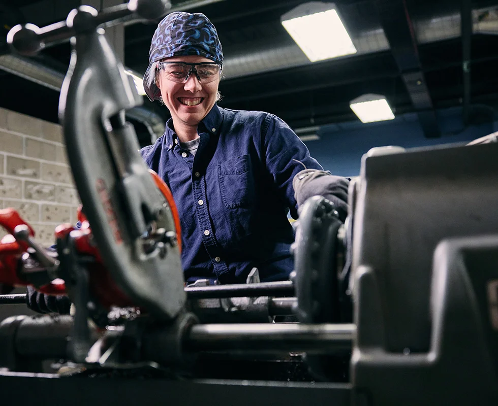 Saint Paul College student wearing safety glasses and protective gear operates pipefitting equipment in a hands-on trades lab.
