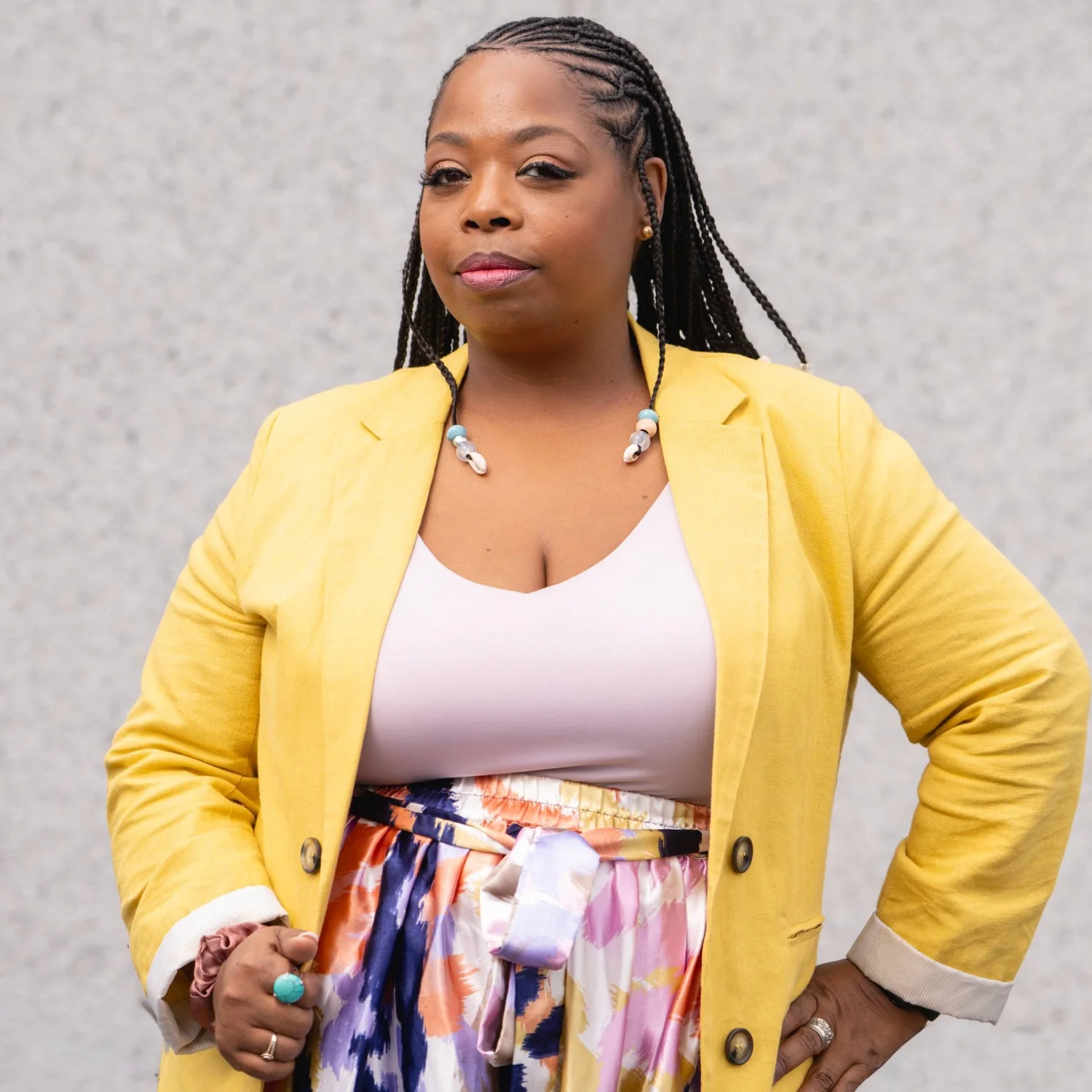 Tameka Jone standing in front on a wall wearing a blazer, skirt, and scoop neck shirt.
