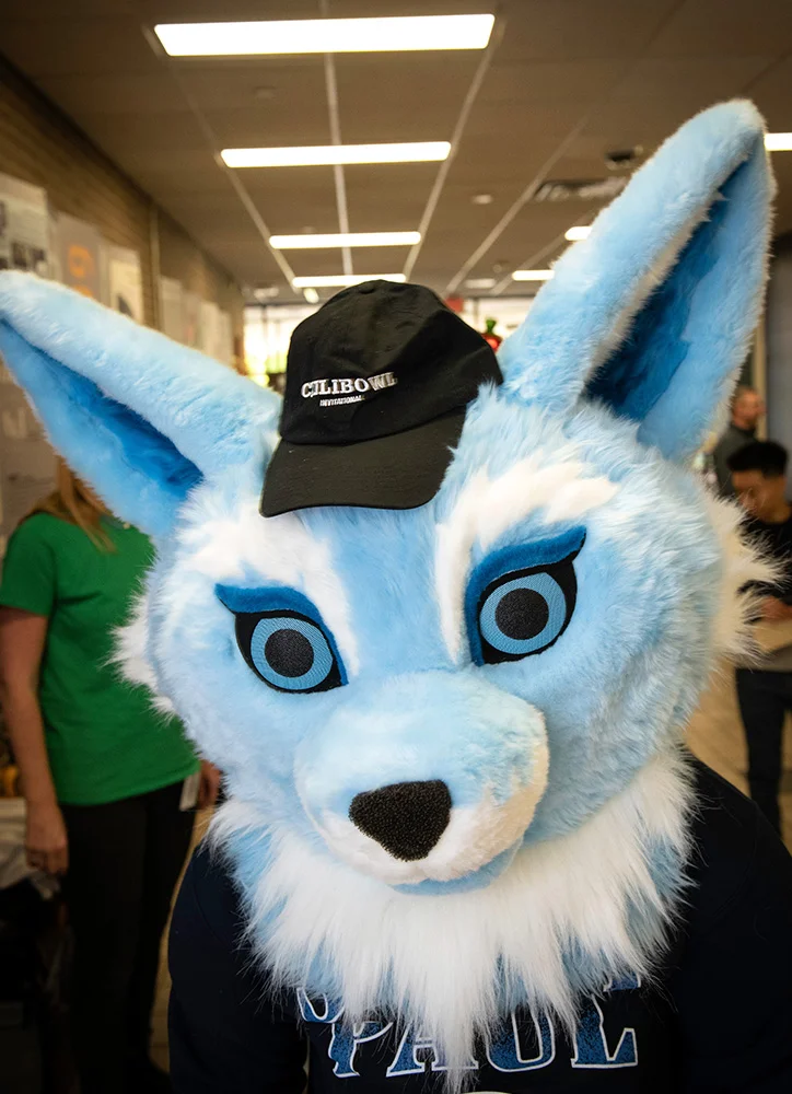 A blue-and-white animal mascot wearing a black Chili Bowl International cap poses indoors at a campus event with attendees in the background.