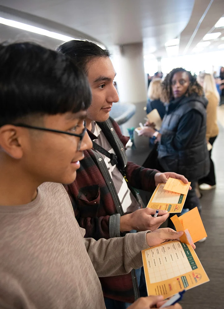 Students hold event cards and write on them while participating in an activity during a busy campus gathering