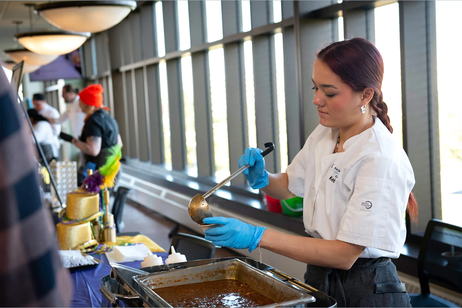Student in a culinary uniform and blue gloves serves chili from a warming tray into a cup at a campus event table decorated with colorful Mardi Gras-themed decorations.